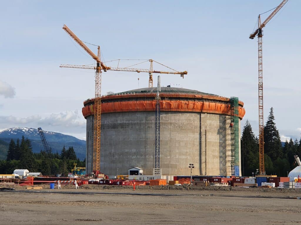 Large circular concrete storage tank under construction, surrounded by cranes, scaffolding, and construction equipment, with forested mountains in the background.