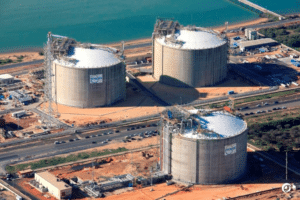 Vue aérienne d’un site industriel côtier avec trois grandes structures cylindriques en béton, des installations techniques et des voies de circulation à proximité de l’eau.