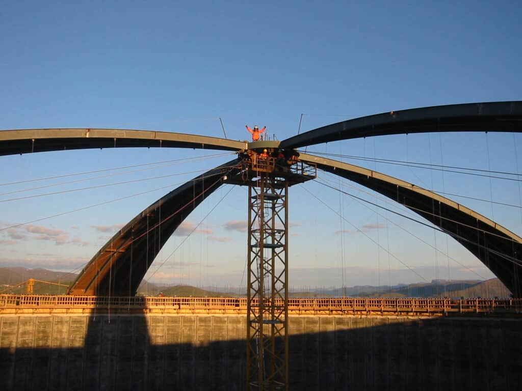Structure métallique arquée en cours de montage, soutenue par un pylône central, avec des câbles et des ouvriers en tenue de sécurité au sommet.