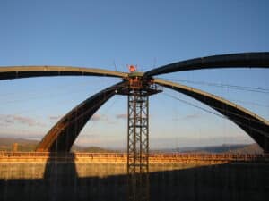 Structure métallique arquée en cours de montage, soutenue par un pylône central, avec des câbles et des ouvriers en tenue de sécurité au sommet.