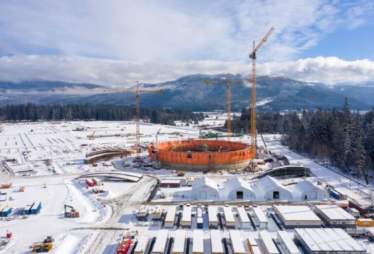 Chantier industriel enneigé avec un grand réservoir circulaire en construction, entouré de grues, de bâtiments temporaires et de montagnes boisées en arrière-plan.