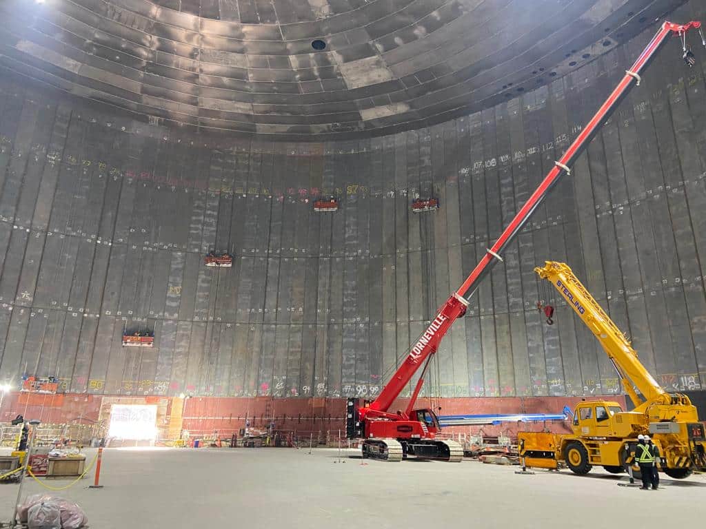 Interior view of a large circular storage tank under construction, showing curved concrete walls, overhead steel panels, and cranes with workers operating on the floor.