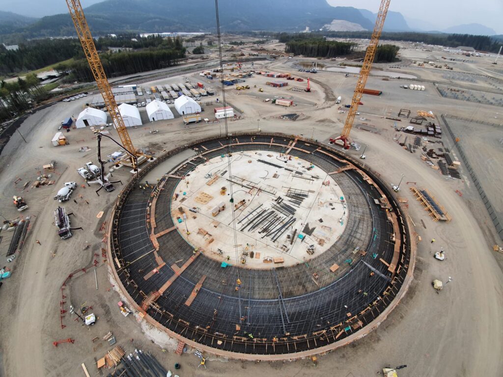 Aerial view of a large circular industrial tank under construction, with reinforcement steel, workers, and two cranes, set within a wide construction site surrounded by roads, equipment, and forested mountains.