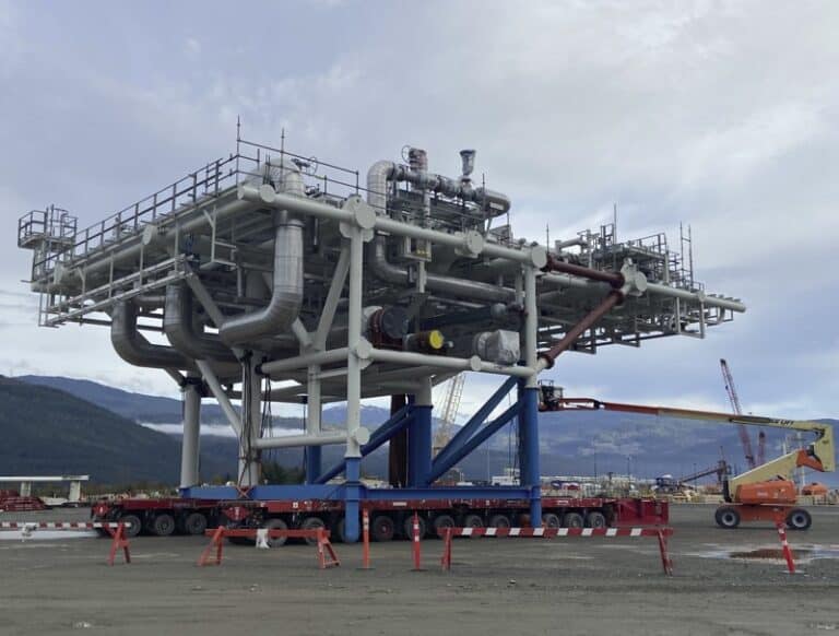 Large prefabricated industrial piping module mounted on a transport platform, with steel pipes, valves, and access platforms, positioned on a construction site with mountains in the background.