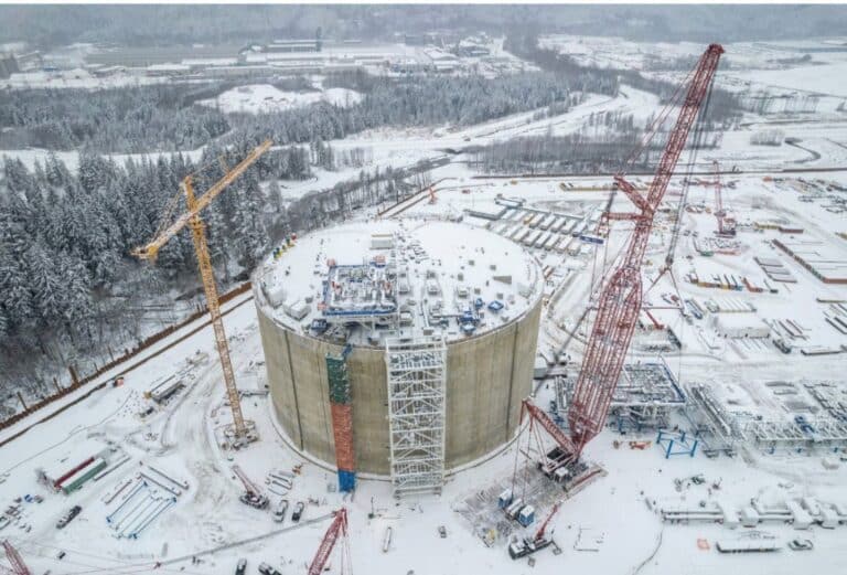 Aerial view of a large circular concrete storage tank under construction in a snow-covered industrial site, with cranes, scaffolding, and equipment around the structure and forested landscape in the background