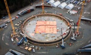 Aerial view of a circular tank foundation under construction, with cranes, formwork, and materials arranged around the concrete base within an active industrial site.